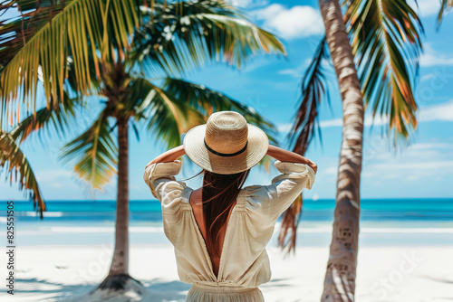 A woman in a casual linen outfit standing with her back facing the camera, holding her hat with two hands and posing for a photo on a white sand beach with beautiful palm trees. The scene exudes an el