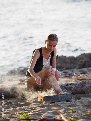 Woman cooking fried chicken on a tropical sandy beach, near the ocean.