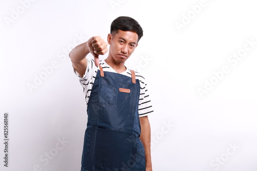 Young Asian man wearing blue appron looking unhappy and angry showing rejection and negative with thumbs down gesture. bad expression photo