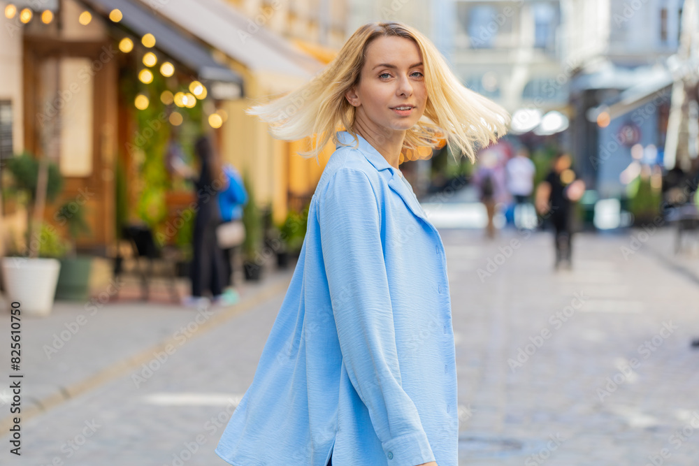 Rear view of young happy blonde woman tourist walking through European street outdoors turns and looking at camera. Back view of lovely pretty girl lady traveler walk in urban city. Town lifestyles Rear view of young happy blonde woman tourist walking through European street outdoors turns and looking at camera. Back view of lovely pretty girl lady traveler walk in urban city. Town lifestyles