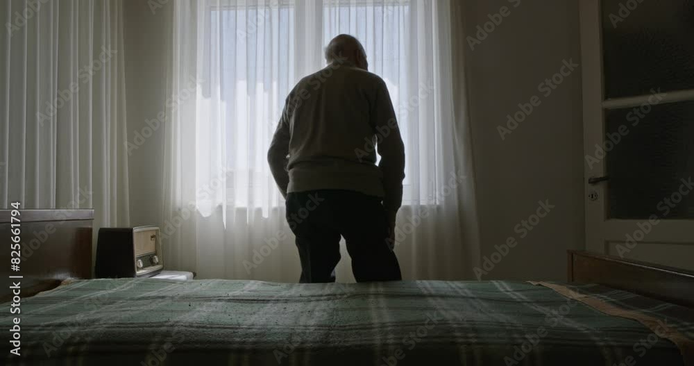 An elderly man sits alone on a bed in a sunlit room, staring out a window with sheer curtains.