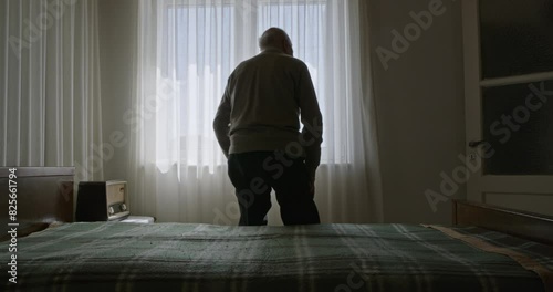 An elderly man sits alone on a bed in a sunlit room, staring out a window with sheer curtains.