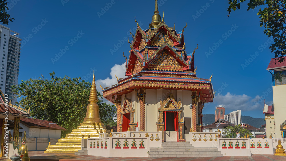 Beautiful Thai Buddhist temple. A building with a roof decorated with ornaments and bas-reliefs. A golden spire against the blue sky. The steps lead to the door. City houses away. Malaysia. Penang. 