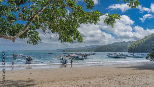 Many Filipino bangka boats are anchored and moored to the shore of the turquoise ocean. The waves are foaming on the beach. Silhouettes of people. Hills against a blue sky and clouds. Green  branches 