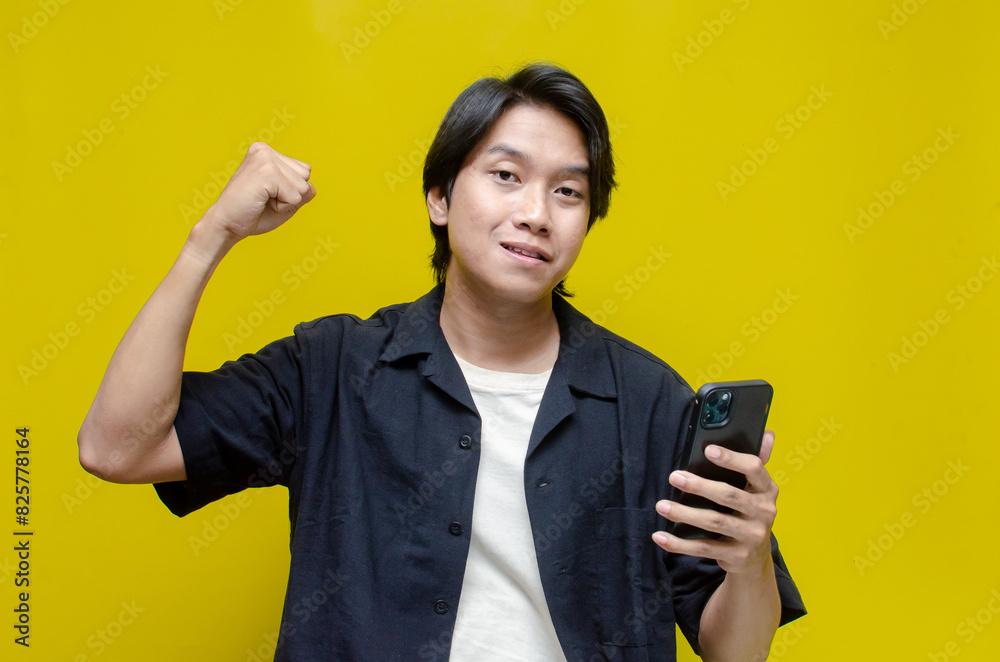 a funny young Asian man holding phone and dancing happily. asian man with joyful expression holding phone and making yes gesture after receiving a good news. a funny young Asian man holding phone and dancing happily. asian man with joyful expression holding phone and making yes gesture after receiving a good news.