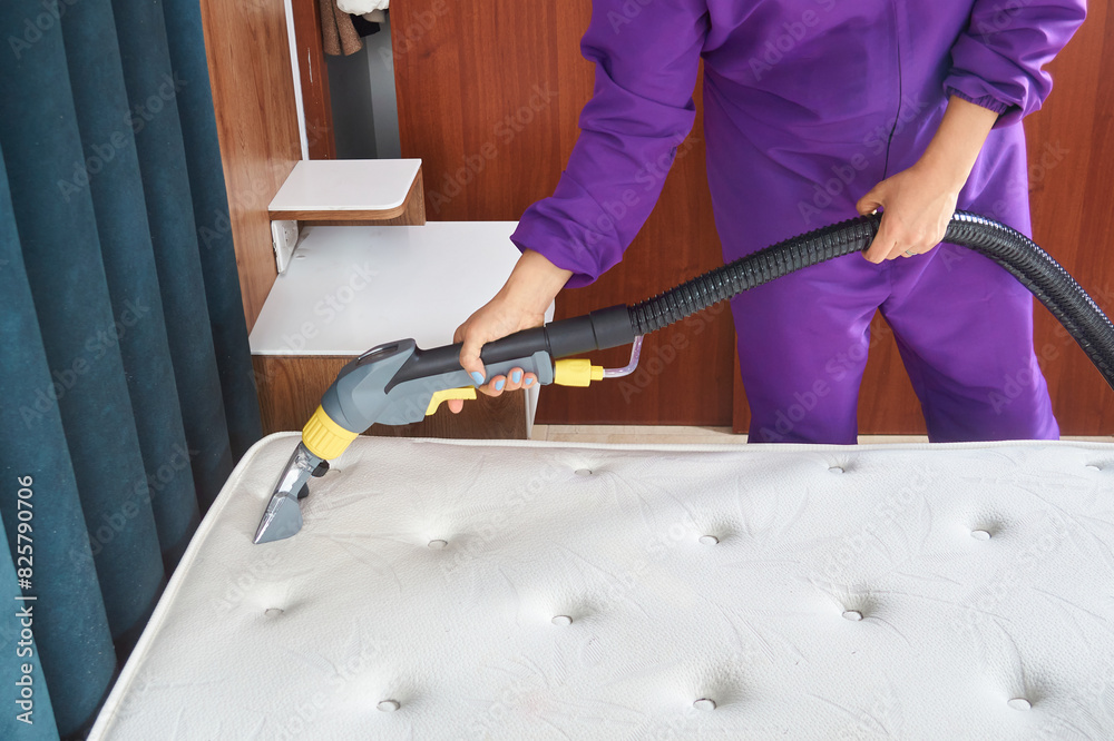 A person in a purple jumpsuit is cleaning a mattress with a steam cleaner