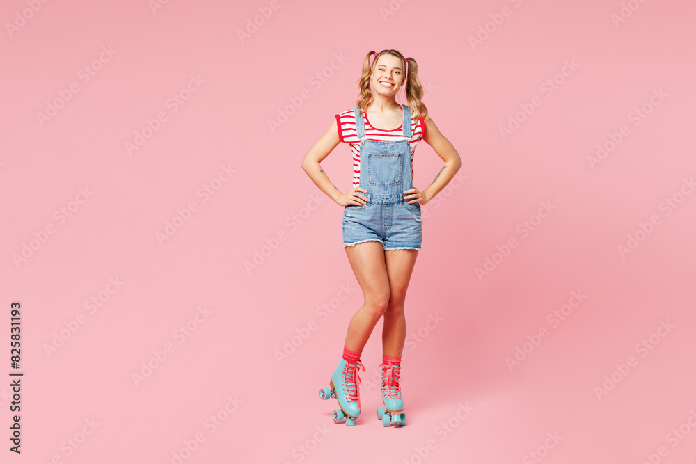 Full body young happy woman wear red t-shirt denim overalls casual clothes blue rollers rollerblading stand akimbo look camera isolated on plain pink background Summer sport lifestyle leisure concept.