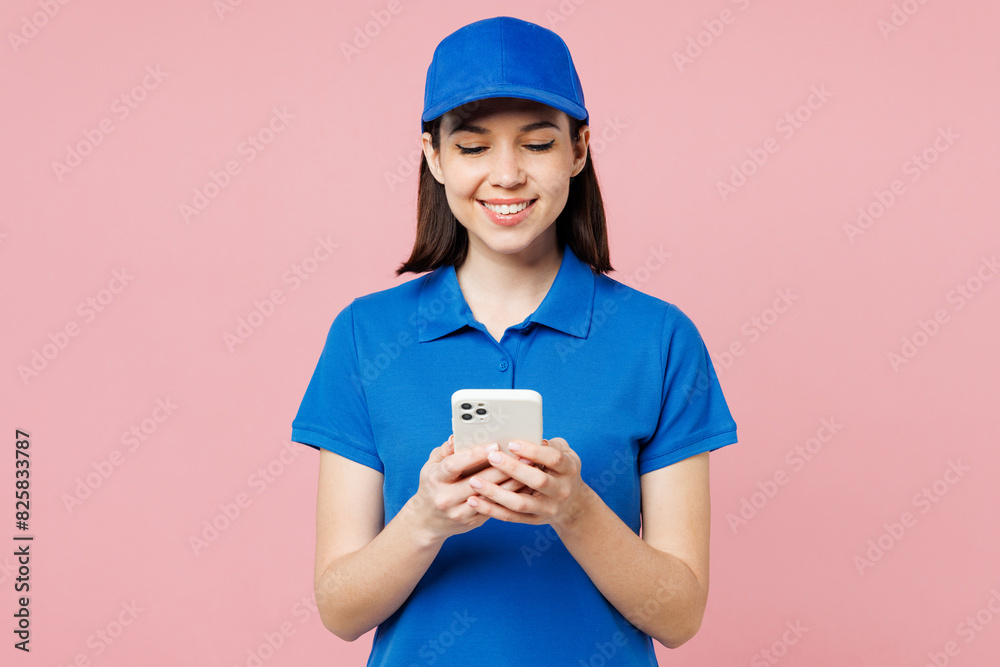 Professional smiling delivery girl employee woman wear blue cap t-shirt uniform workwear work as dealer courier hold in hand use mobile cell phone isolated on plain pink background. Service concept. Professional smiling delivery girl employee woman wear blue cap t-shirt uniform workwear work as dealer courier hold in hand use mobile cell phone isolated on plain pink background. Service concept.
