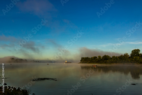 Fishermen on boats early in the morning at dawn in the fog at golden hour catch fish on the Oka River, Russia. Barge, tugboat, green grass and trees, a river bend against a clear light sky in summer.