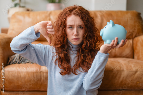 Photography unhappy redhead woman with grimace of displeasure holding piggy bank and giving thumb down saving money for rainy days investments, deposits