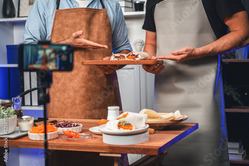 Cropped view of chef influencers presenting fresh salad roll on cooking step by step, streaming via smartphone on social media live channel, showing ingredients vegetable soft tortilla wrap. Sellable. photo