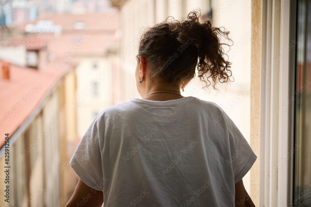 Rear view. Young woman in white t-shirt, enjoying the morning of her holidays, contemplating through the window the beautiful touristic city of Como with red roof tiles of cozy houses Rear view. Young woman in white t-shirt, enjoying the morning of her holidays, contemplating through the window the beautiful touristic city of Como with red roof tiles of cozy houses