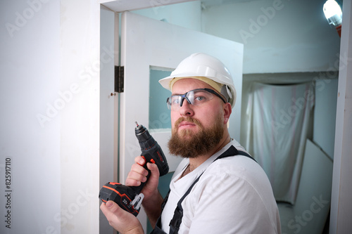 Construction worker with a screwdriver installing a door. Accessories for assembling, install furniture, repair home. Man dressed in work attire, helmet and protective glasses. Home renovation concept