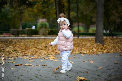 Small pretty girl with baby carriage walking in the park full of colorful leaves on the ground. Cute baby girl in the park. Child carrying leaves in the park