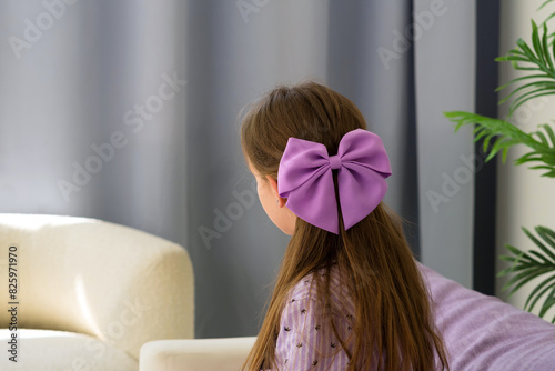 Girl with purple bow in hair sitting by window. (ID: 825971970)