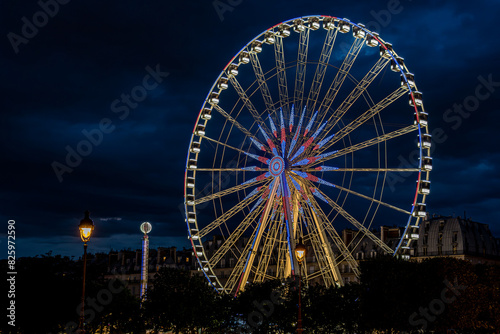 Ferris wheel illuminated with the colors of the French flag red  white and blue  over a cloudy sky and with the typical roofs of Paris in the background.