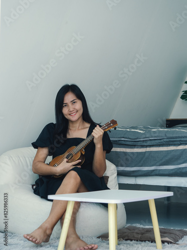 Asian young woman wearing black dress, bright smiling face Sitting and playing the ukulele On a pillow sitting in white room