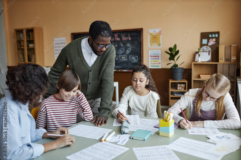 Obraz premium Young African American male teacher of English standing at table in classroom working with multi-ethnic group of students