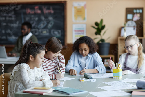 Group of four ethnically diverse boys and girls sitting at table in classroom doing exercises while their teacher using laptop in background