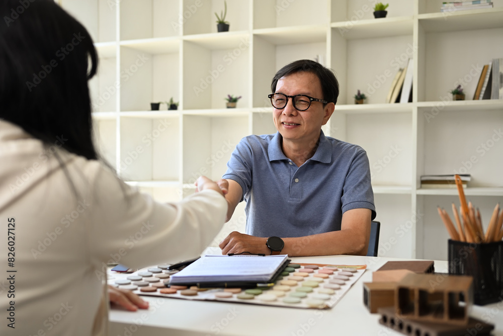 Smiling senior male interior designer shaking hands with his client after signing contract at office Smiling senior male interior designer shaking hands with his client after signing contract at office