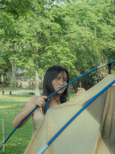Young Asian woman setting up a tent for camping Amidst green nature, The background lined with trees. Holiday activities