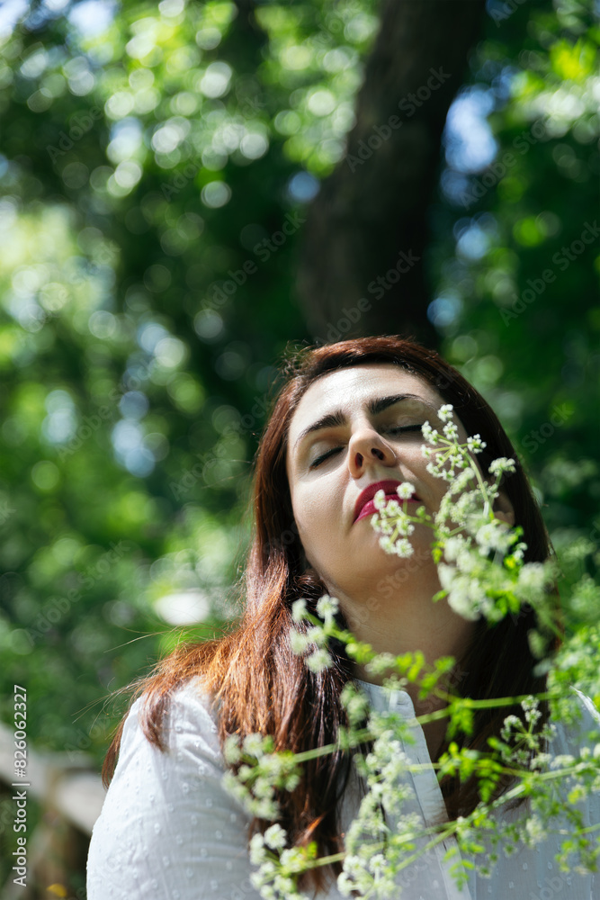 Portrait of a beautiful 45 years old woman with closed eyes relaxing and calm among flowers with the green background of the foliage of a park, on a sunny day. Concept of maturity and well-being Portrait of a beautiful 45 years old woman with closed eyes relaxing and calm among flowers with the green background of the foliage of a park, on a sunny day. Concept of maturity and well-being