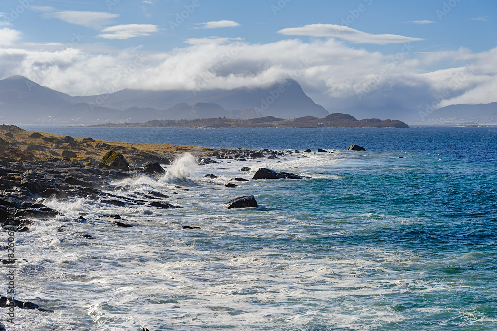 Waves crash against the rocky shore as clouds hover above distant mountains. The serene coastline contrasts with the power of the ocean under a bright blue sky. Waves crash against the rocky shore as clouds hover above distant mountains. The serene coastline contrasts with the power of the ocean under a bright blue sky.