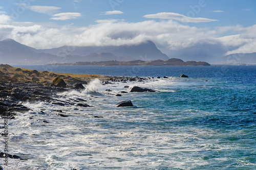 Waves crash against the rocky shore as clouds hover above distant mountains. The serene coastline contrasts with the power of the ocean under a bright blue sky.