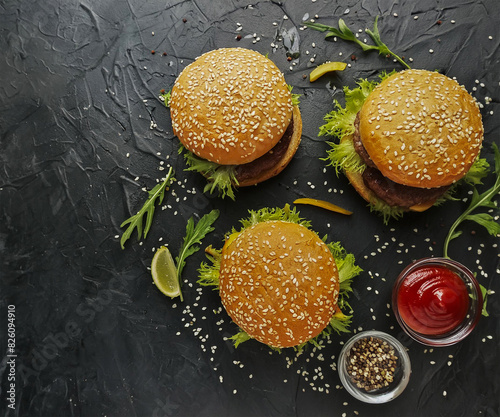 Three sesame seed buns hold juicy burgers layered with lettuce  onion  tomato  melted cheese  and beef patties on a dark surface. Surrounding greens  sesame seeds  ketchup  and mustard add flavor.