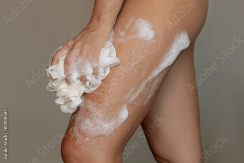 Cropped shot of a woman massaging her thighs with a rose washcloth with abundant foam on a dark gray background. Girl washing her body in the shower with gel. Cosmetology, peeling and spa cosmetics