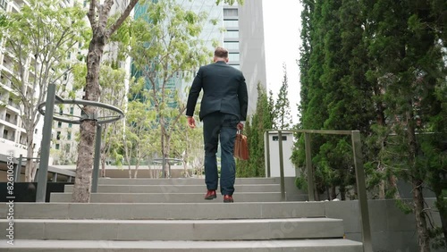 Back view of caucasian manager with formal suit walking up stairs while holding his suitcase in green city. Professional business man going to workiplace while climb stair in eco urban city. Urbane.. photo