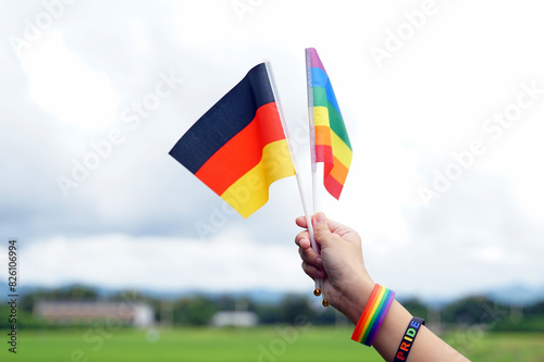 hand wearing a rainbow wristband holds the flag of Germany and the rainbow flag, a symbol of LGBT pride, holding the flag high against the background of the sky.