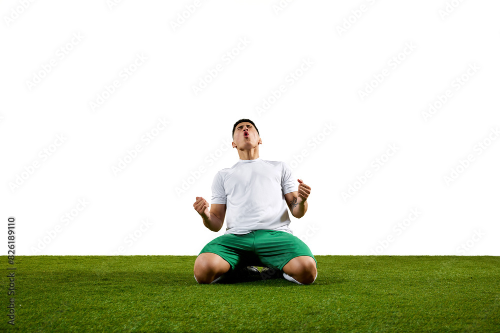 High-energy image of footballer kneeling on grassland with fists raised in celebration against white studio background. Concept of professionals sport, competition, tournament, energy, action. Ad High-energy image of footballer kneeling on grassland with fists raised in celebration against white studio background. Concept of professionals sport, competition, tournament, energy, action. Ad