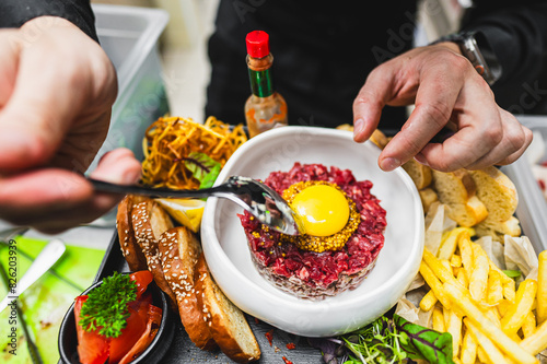 Close-up of a hand holding a fork over a plate of steak tartare topped with a raw egg yolk, surrounded by fries, bread, and greens
