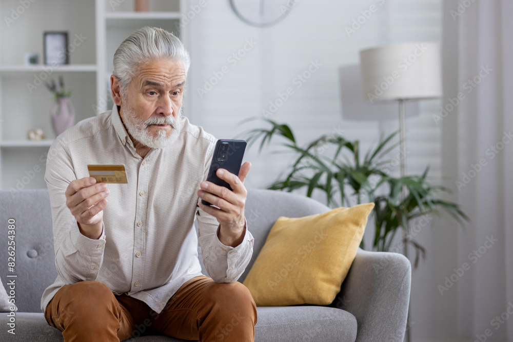 A senior man in a white shirt with gray hair and a beard is sitting on the sofa at home, holding a bank credit card in his hand and looking worriedly at the mobile phone screen A senior man in a white shirt with gray hair and a beard is sitting on the sofa at home, holding a bank credit card in his hand and looking worriedly at the mobile phone screen