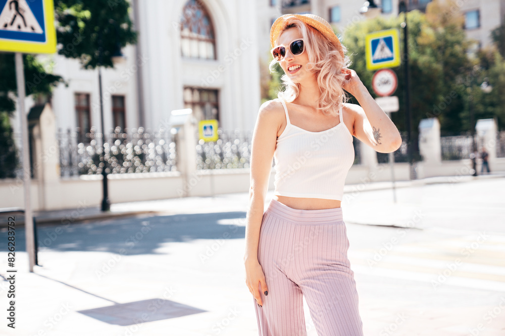 Fototapeta premium Young beautiful smiling blond hipster woman in trendy summer clothes. Carefree female posing in the street at sunny day. Positive model outdoors at sunset. Cheerful and happy in hat, sunglasses
