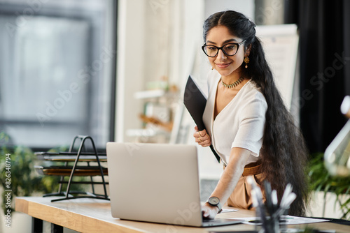 A stylish indian woman in glasses works diligently on a laptop in an office setting.