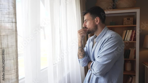Young sad and depressed man feeling regret and anxiety, standing near window looking through. Mental burnout after hard work and hours of insomnia. Relationship difficulties desperation and depression