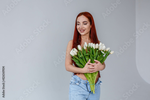Obraz na płótnie Red-haired girl in jeans with white tulips posing on a white background
w salonie