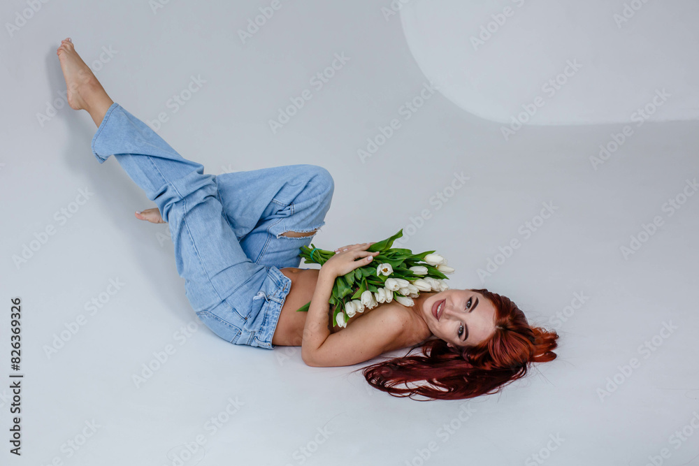 Red-haired girl in jeans with white tulips posing while lying on a white background