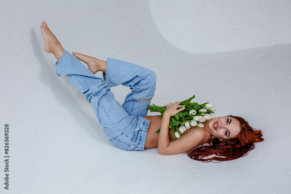 Red-haired girl in jeans with white tulips posing while lying on a white background