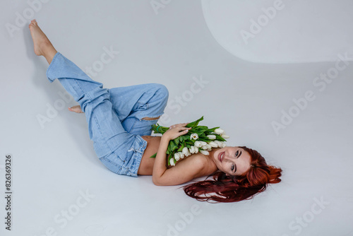 Obraz na płótnie Red-haired girl in jeans with white tulips posing while lying on a white background
w salonie