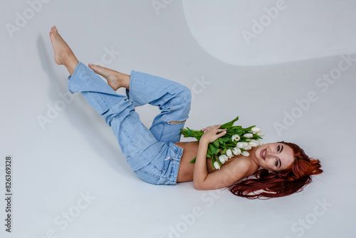 Obraz na płótnie Red-haired girl in jeans with white tulips posing while lying on a white background
w salonie