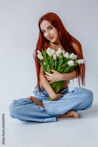 Red-haired girl in jeans with white tulips posing on a white background
