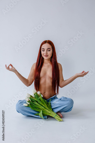 Red-haired girl in jeans with white tulips doing yoga while sitting on a white background