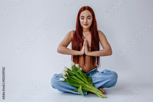 Red-haired girl in jeans with white tulips doing yoga while sitting on a white background