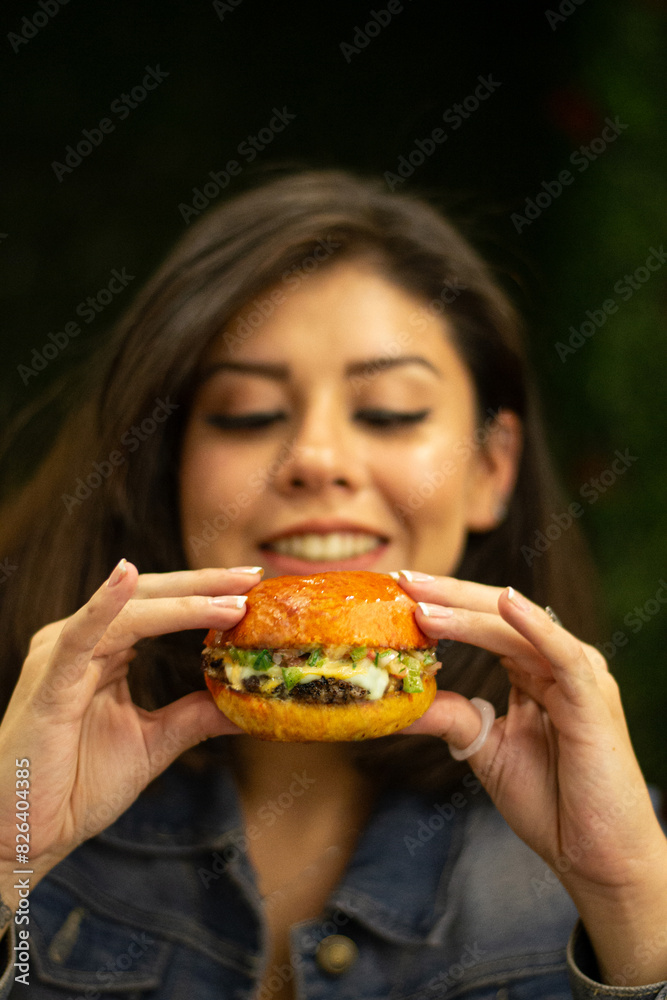 A hispanic woman is holding a burger in her hands and smiling, enjoying her meal, the burger is a large. A hispanic woman is holding a burger in her hands and smiling, enjoying her meal, the burger is a large.