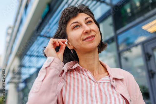 Successful businesswoman using wireless headphones outdoors near her office, portrait of young woman in fashionable clothes, professional manager working on mobile device
