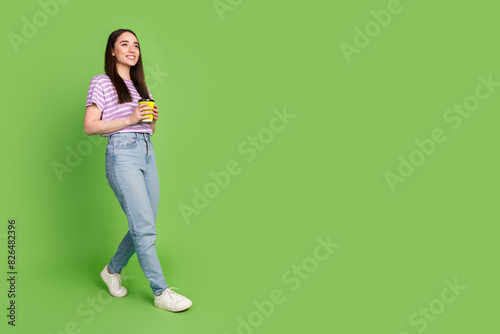 Full body photo of attractive young woman walk hold paper cup dressed stylish striped violet clothes isolated on green color background © deagreez