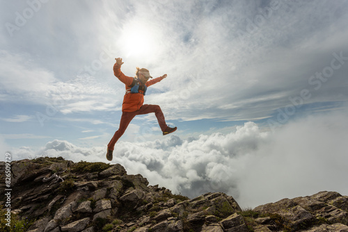 A woman is jumping in the air on a mountain. The sky is cloudy and the sun is shining